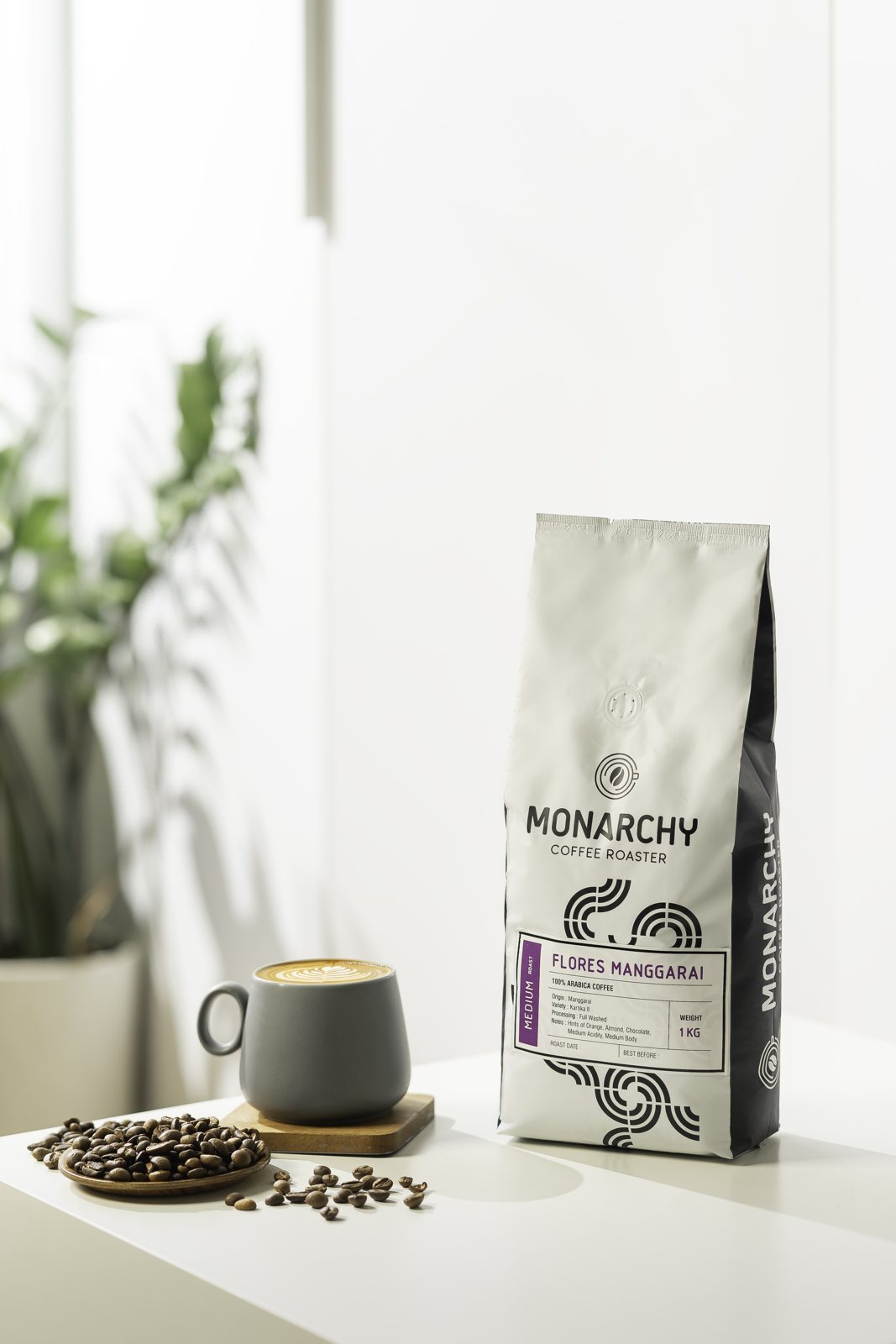 Kemasan kopi Monarchy putih di samping secangkir kopi dan biji kopi