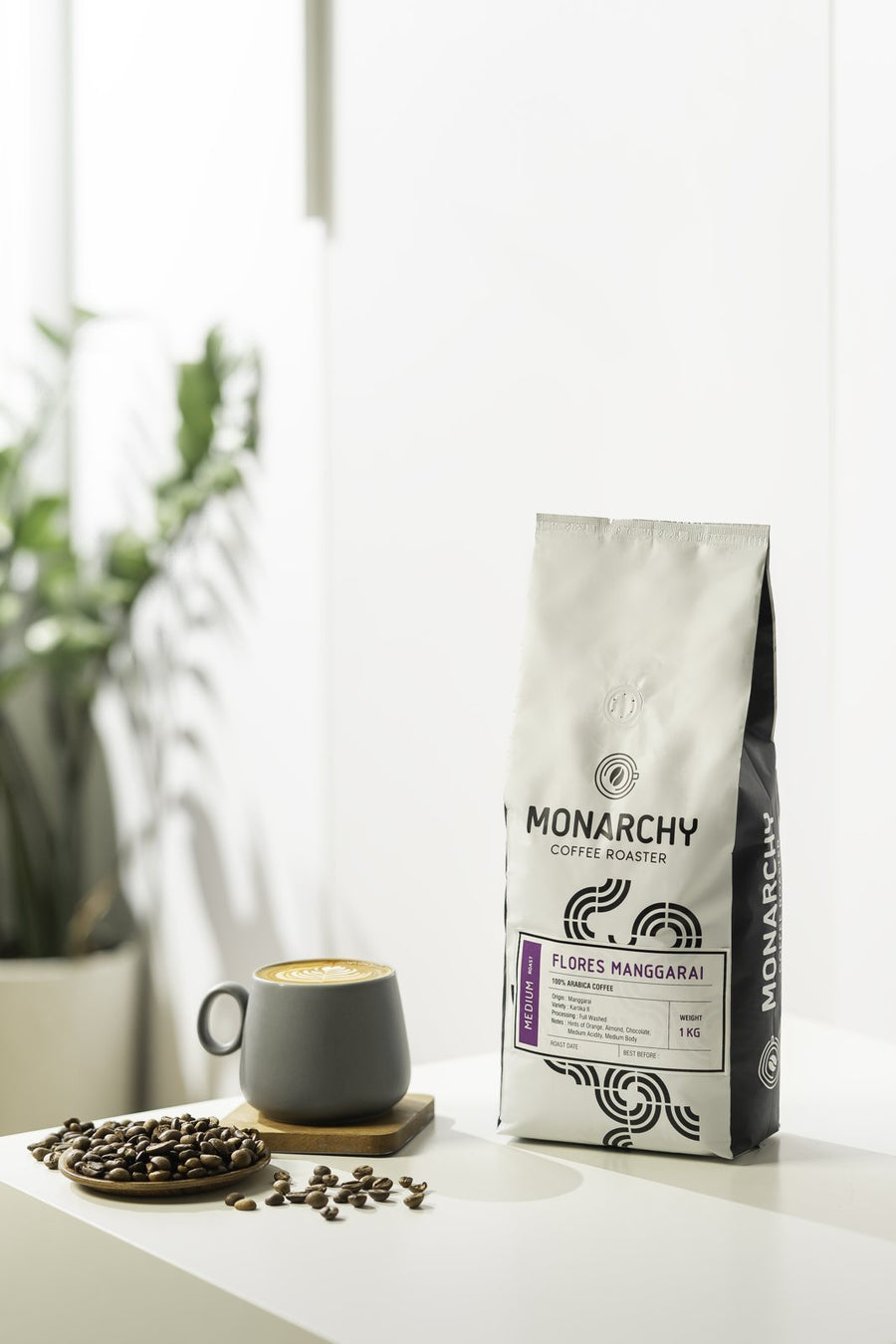 Kemasan kopi Monarchy putih di samping secangkir kopi dan biji kopi