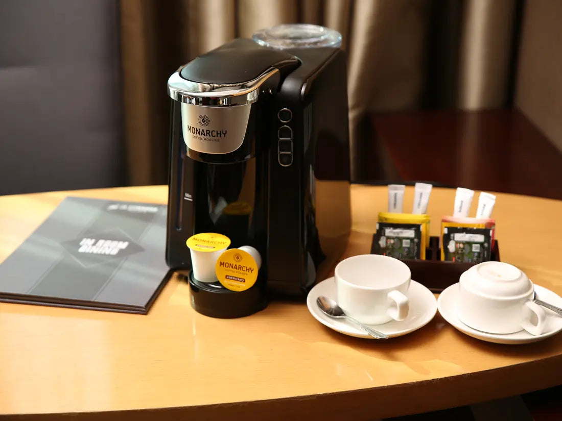 Mesin kopi kapsul Monarchy, kapsul, dan perlengkapan kopi di meja kamar hotel